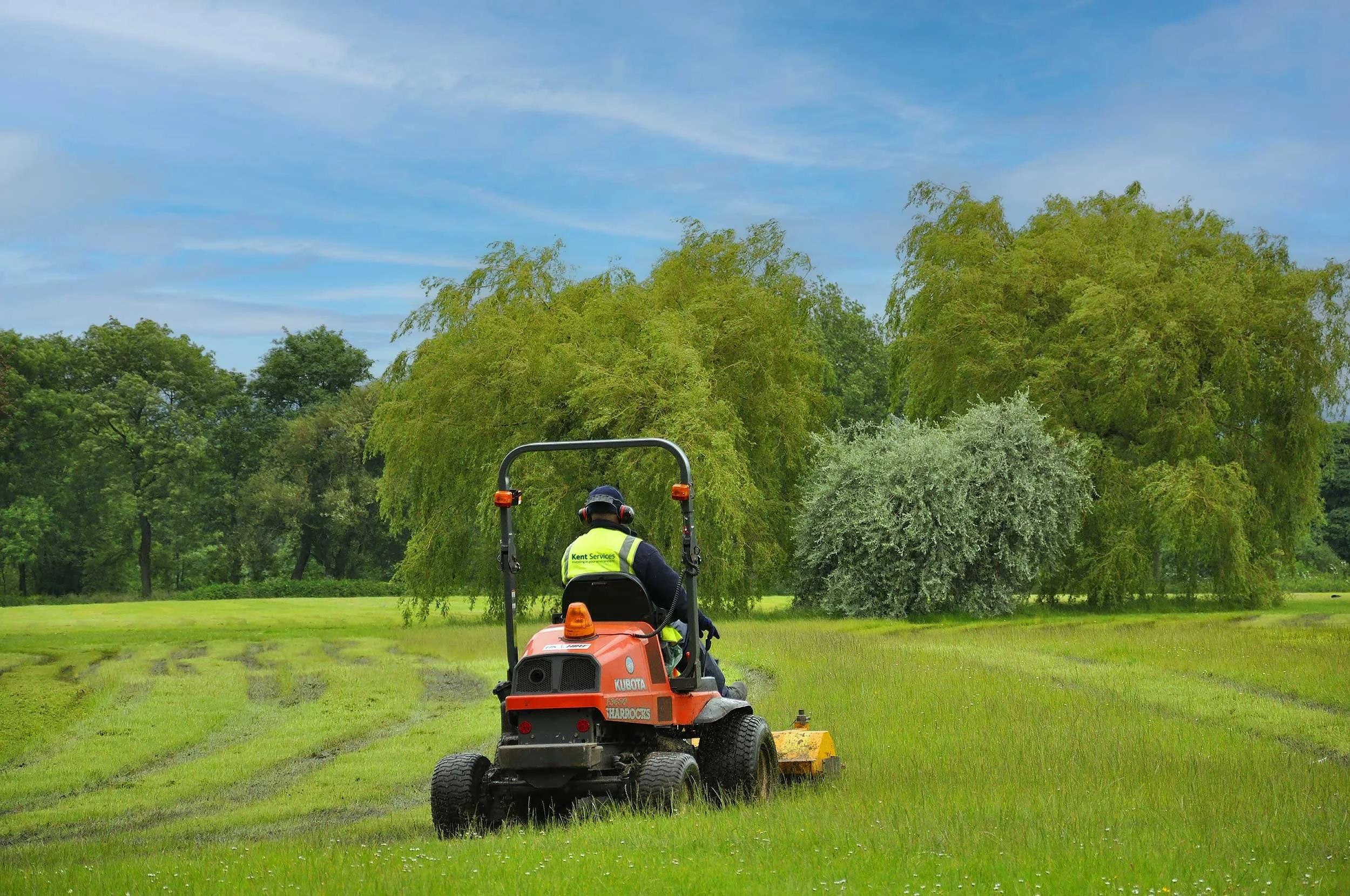 Man operating a red riding mower in a green field with trees and a blue sky with clouds.
