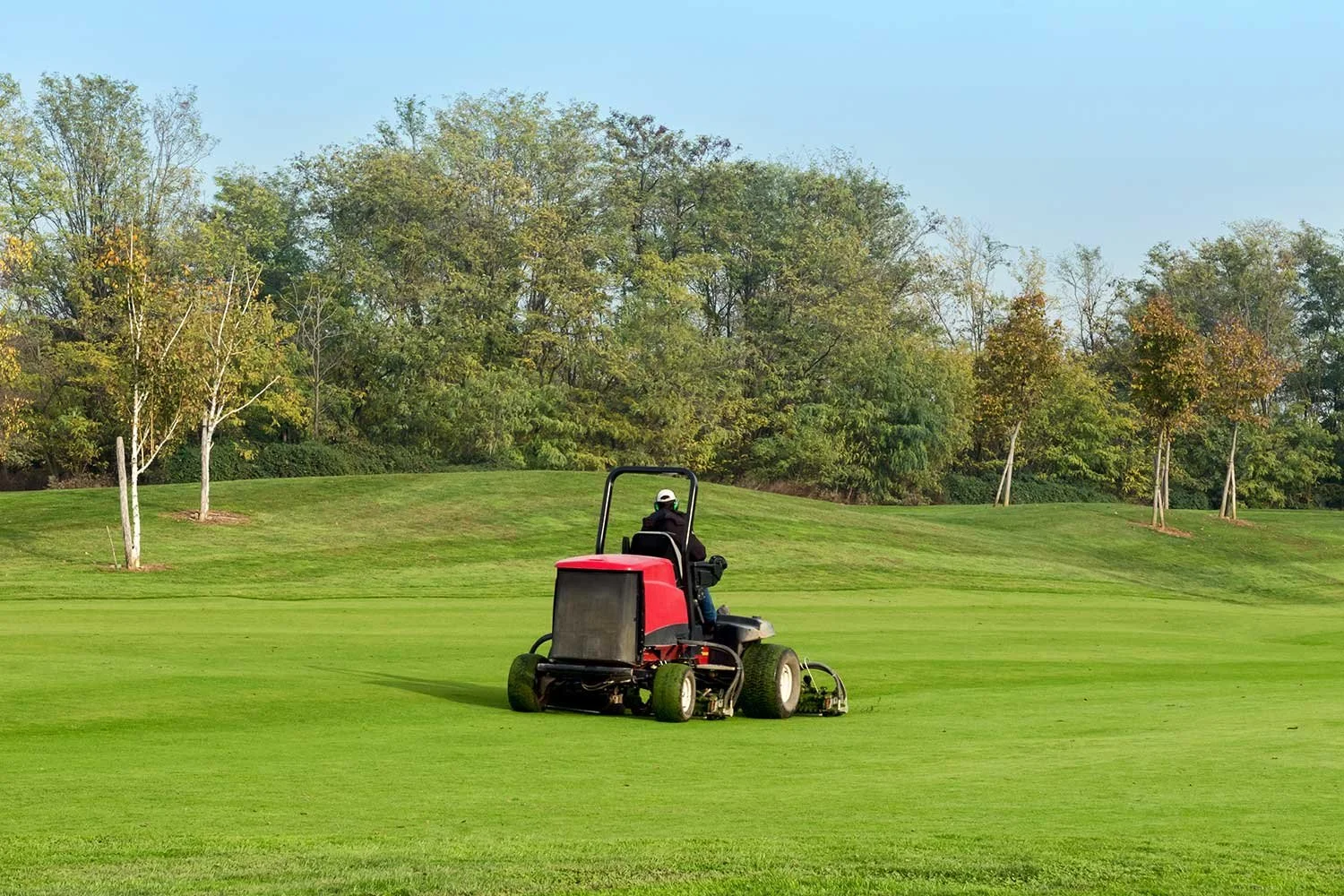 A person operating a small orange lawn tractor on a grassy field, surrounded by trees and a blue sky with some clouds.