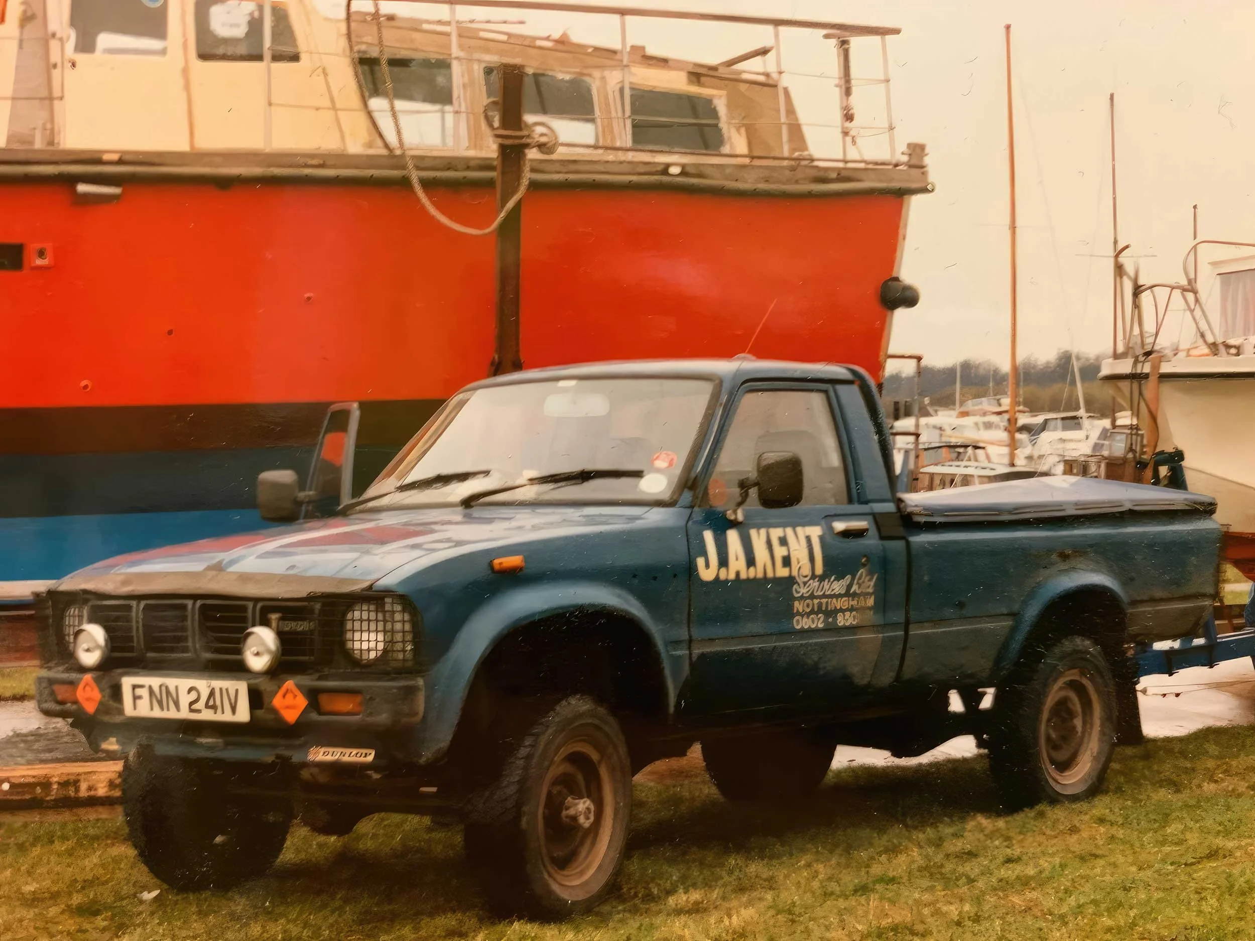 A small blue utility vehicle with the word 'JAKERT' on the door, parked in front of a large red boat with a black stripe, at a marina or boatyard with several boats and masts in the background.