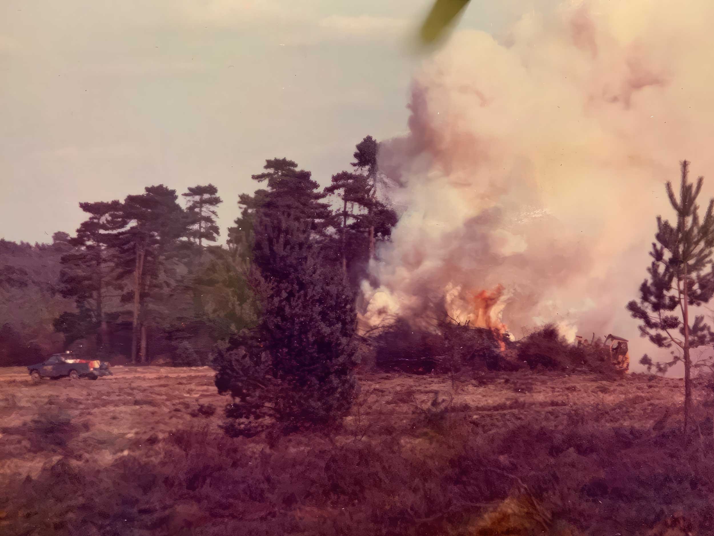 A forest fire with large flames and thick smoke rising over trees in a forested area, with a vehicle on the ground nearby.