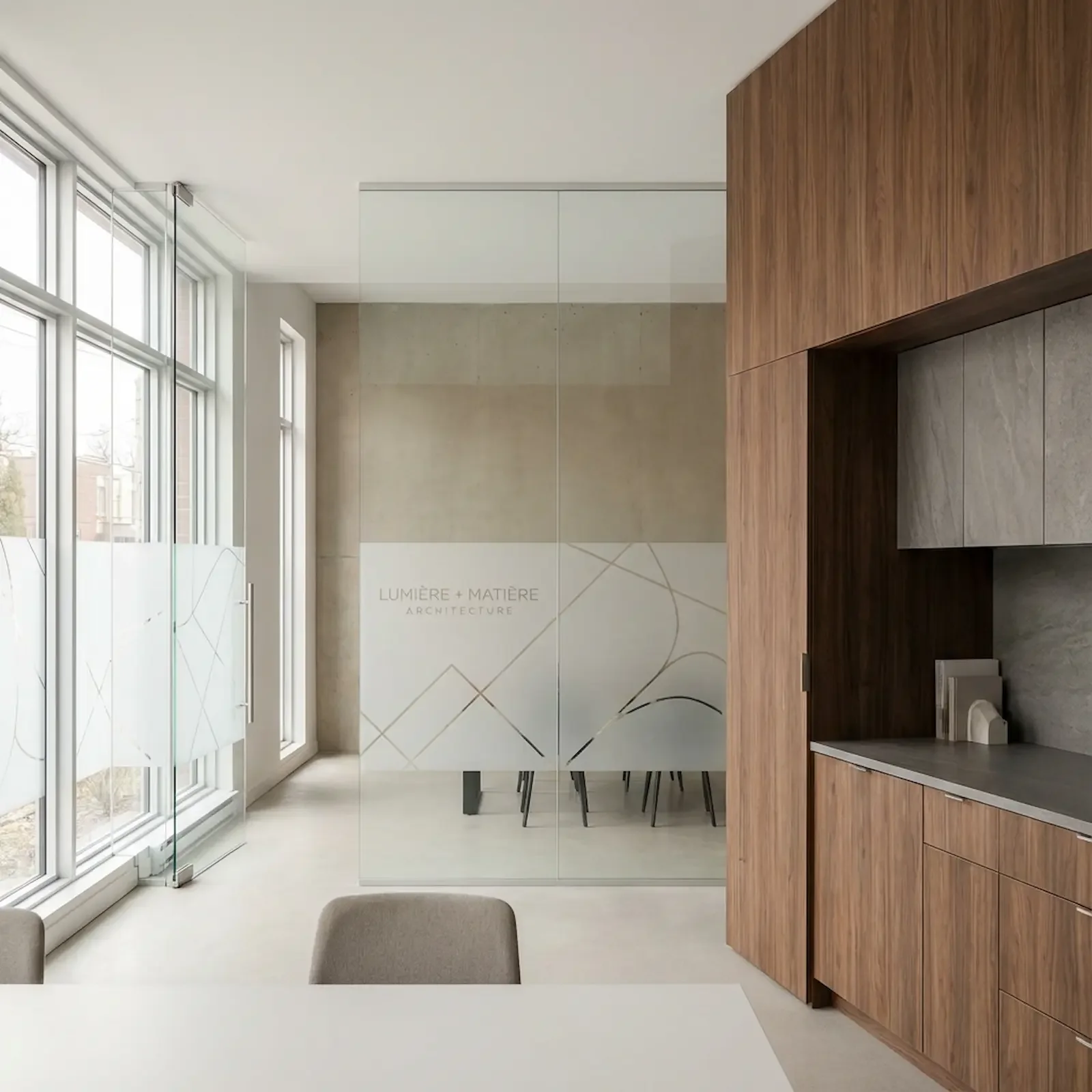 Intérieur moderne avec grandes fenêtres, mur en béton, et meubles en bois. Mur en verre avec inscription "LUMIÈRE + MATIÈRE ARCHITECTURE".