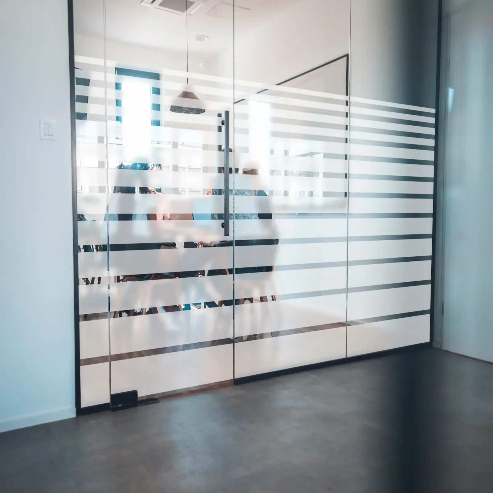 Une porte en verre avec des rayures opaques, derrière laquelle on voit deux personnes en réunion dans une salle de conférence moderne.