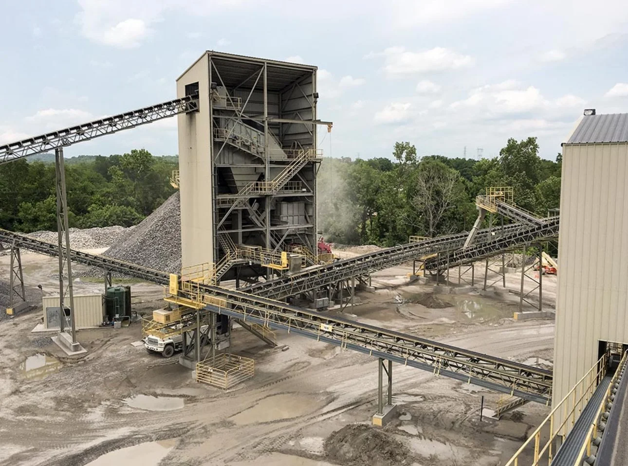 Industrial gravel processing plant with conveyor belts, piles of gravel, and a large central structure, set against a backdrop of green trees and cloudy sky.