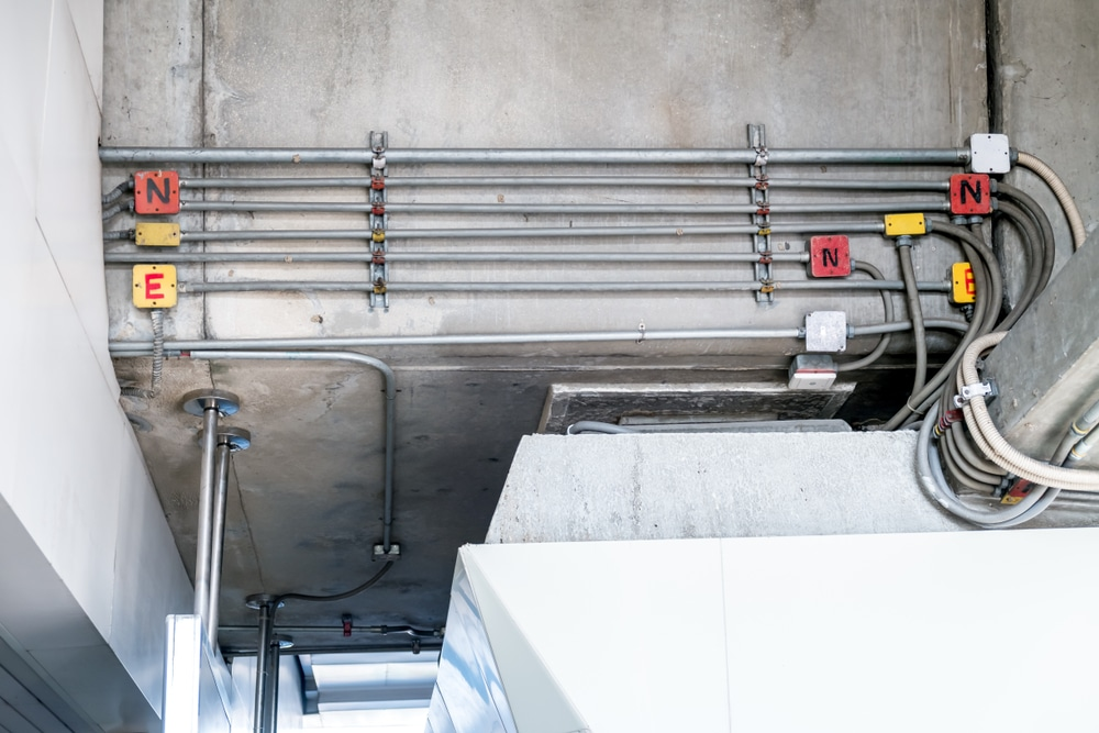 Electrical conduit pipes and cables attached to a concrete ceiling, with red and yellow labeled blocks marked 'N' and 'E'.