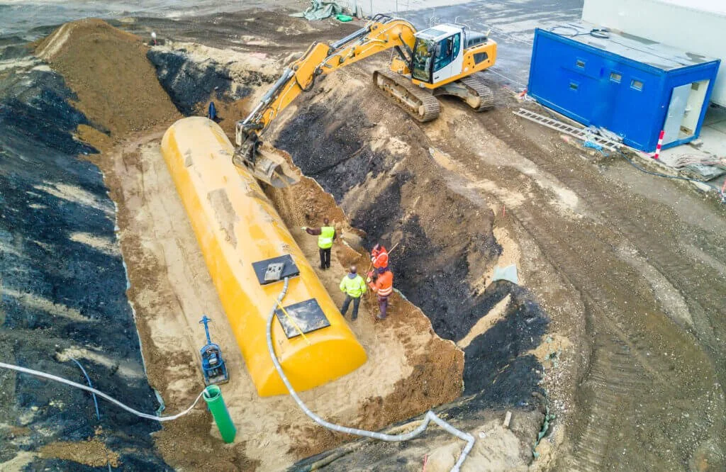 Construction site with an excavator loading dirt into a large yellow tank, three workers in safety vests and helmets near the tank, and a blue portable toilet on the right.