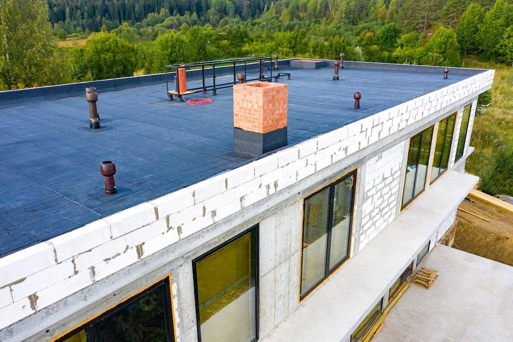 Construction site of a building with a flat roof, brick chimney, vents, and large windows, surrounded by greenery.
