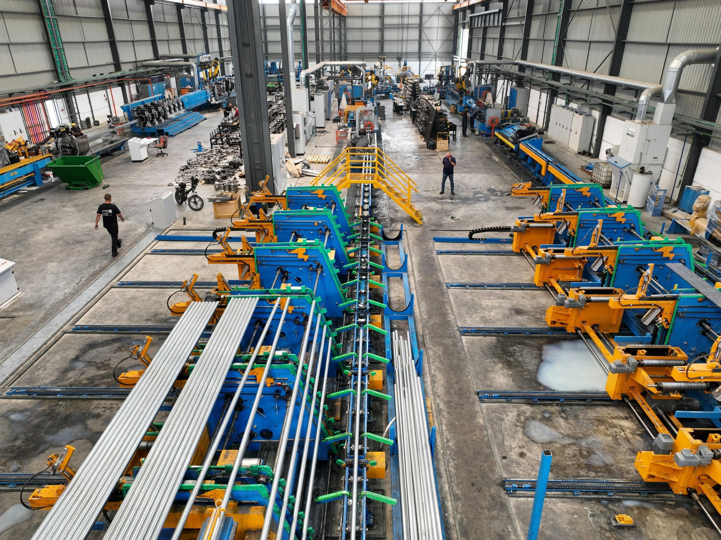 Inside a large industrial factory with various manufacturing machines, metal pipes, and assembly lines. Two workers are present, one walking and one standing, amidst equipment and materials.