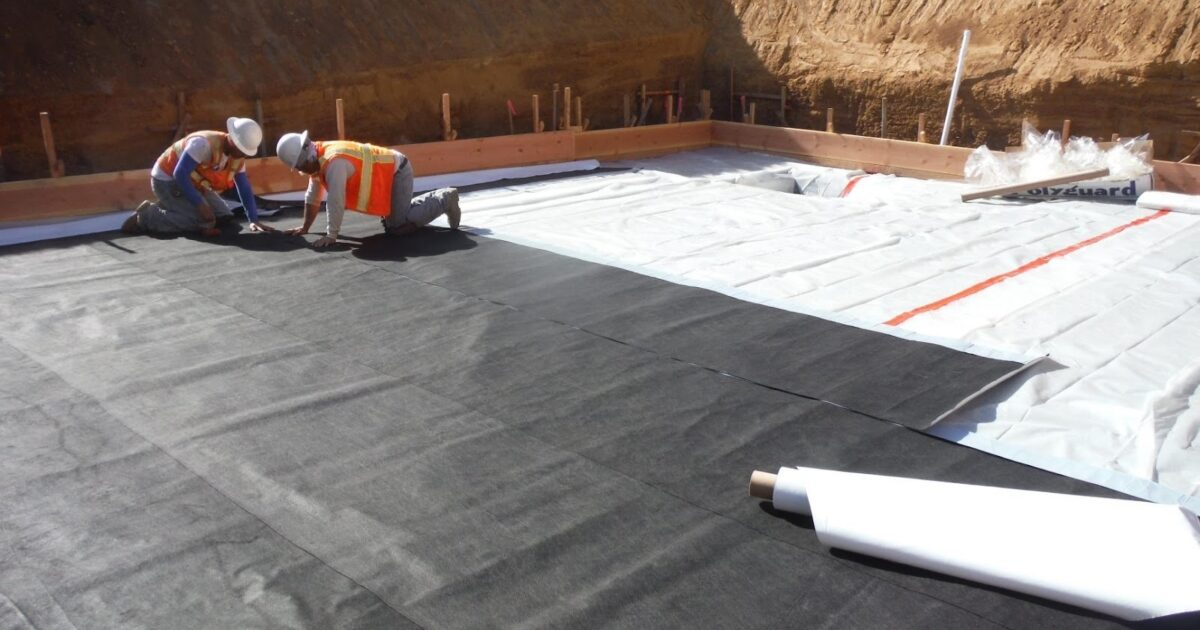 Two construction workers wearing hard hats and safety vests installing a black waterproof membrane on a flat roof at a construction site.
