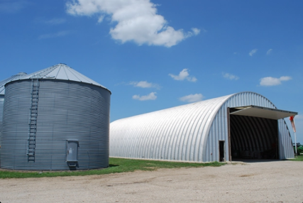 A rural farm scene featuring a large silver grain silo and a white arched metal barn on a grassy area under a blue sky with scattered clouds.