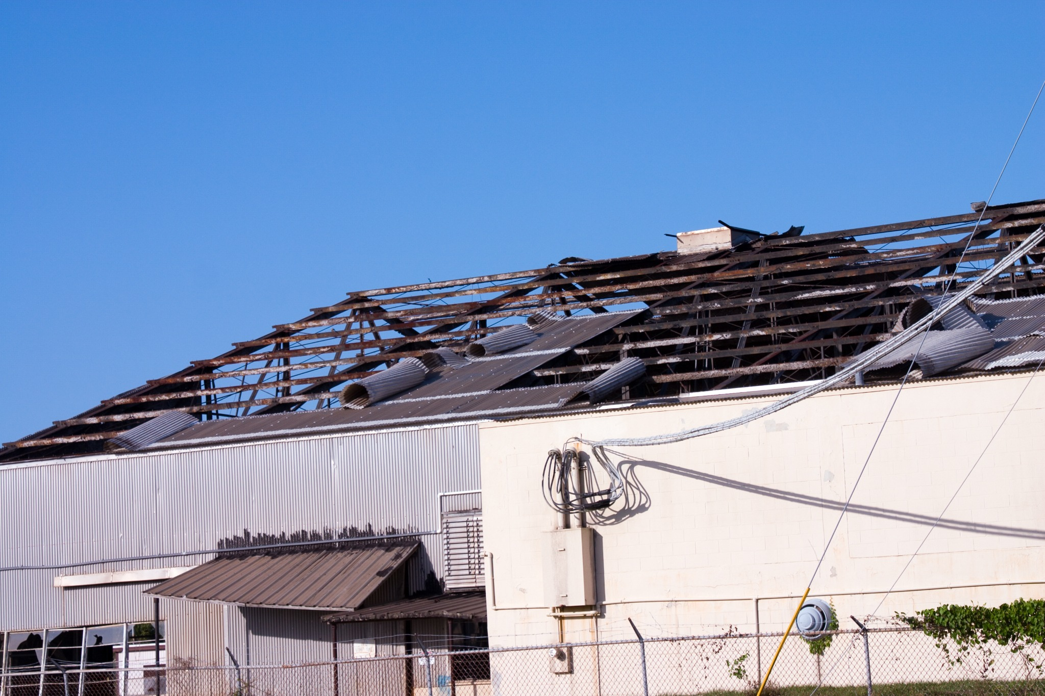 Part of a building's roof with damage, missing shingles, and rusted metal roofing. Clear blue sky background.
