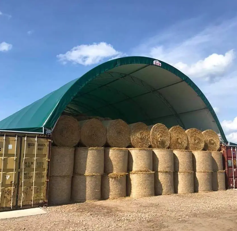 Stacked hay bales inside a green farm storage shelter with open front, blue sky and clouds in the background.
