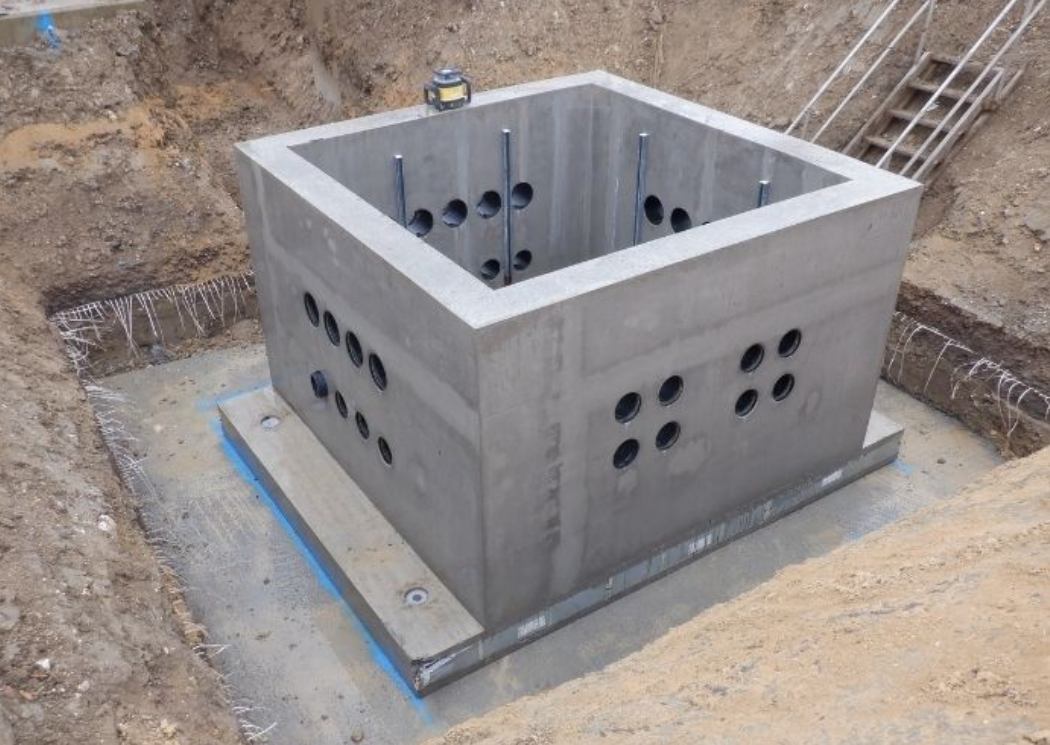 A concrete foundation structure with multiple black pipes, installed in a dug-out area at a construction site.