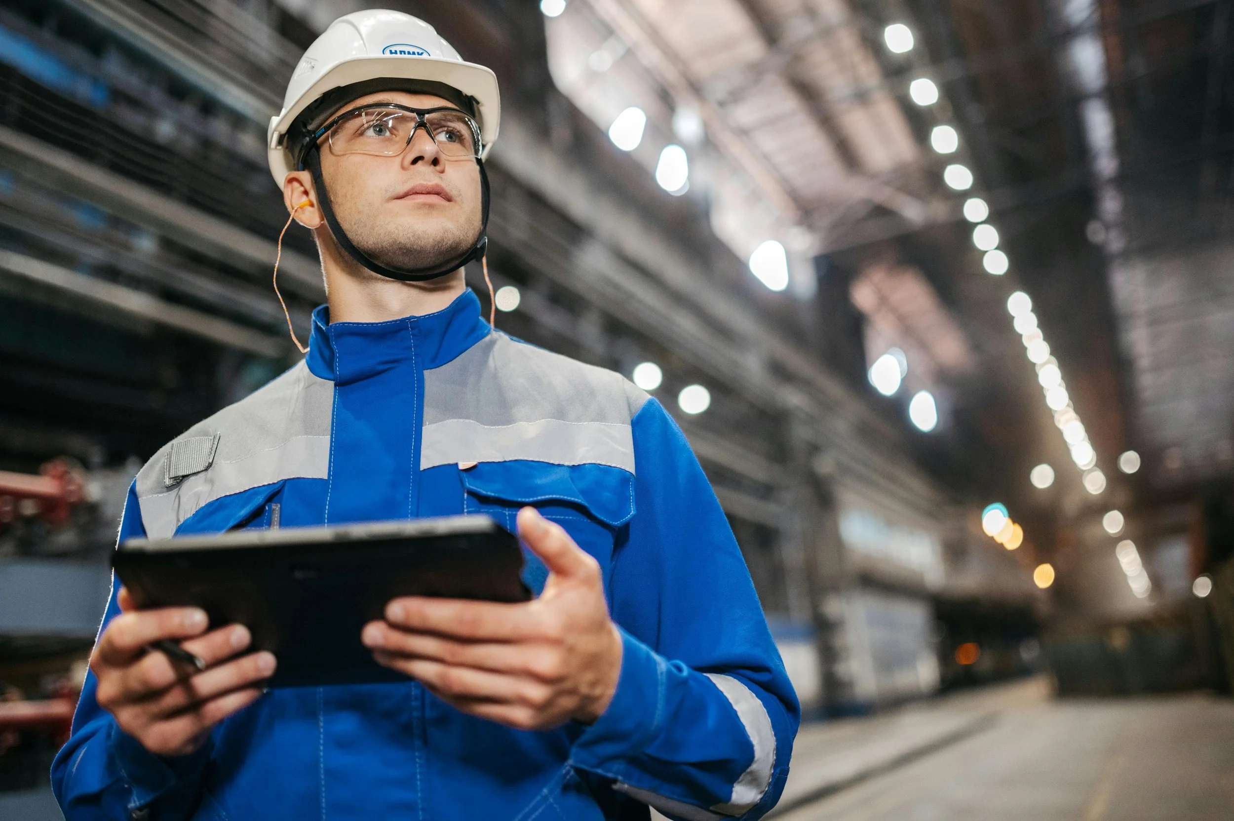 A male industrial worker wearing a safety helmet and glasses, holding a digital tablet inside a factory or warehouse, with blurred background of work environment.