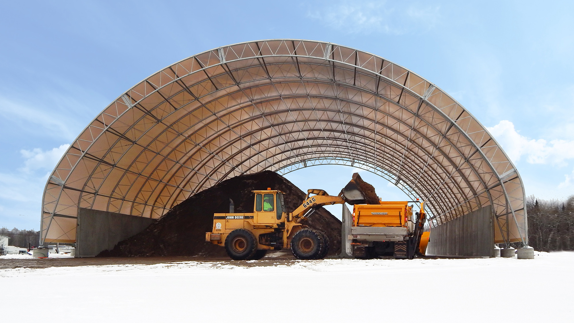 A construction site with a large yellow John Deere front loader moving dark soil into a dump truck inside a semi-circular warehouse with a metal framework, snow on the ground, and a clear blue sky.