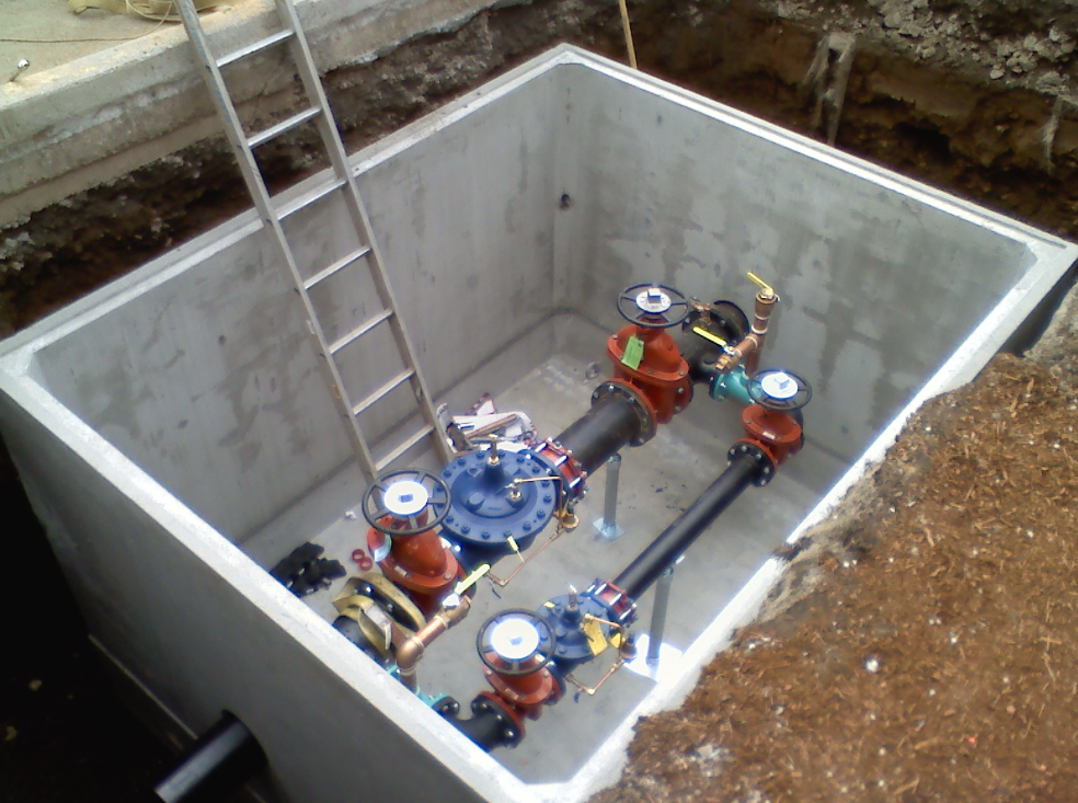 View of an underground water or utility service vault containing water meters, valves, pipes, and a ladder inside a concrete enclosure.