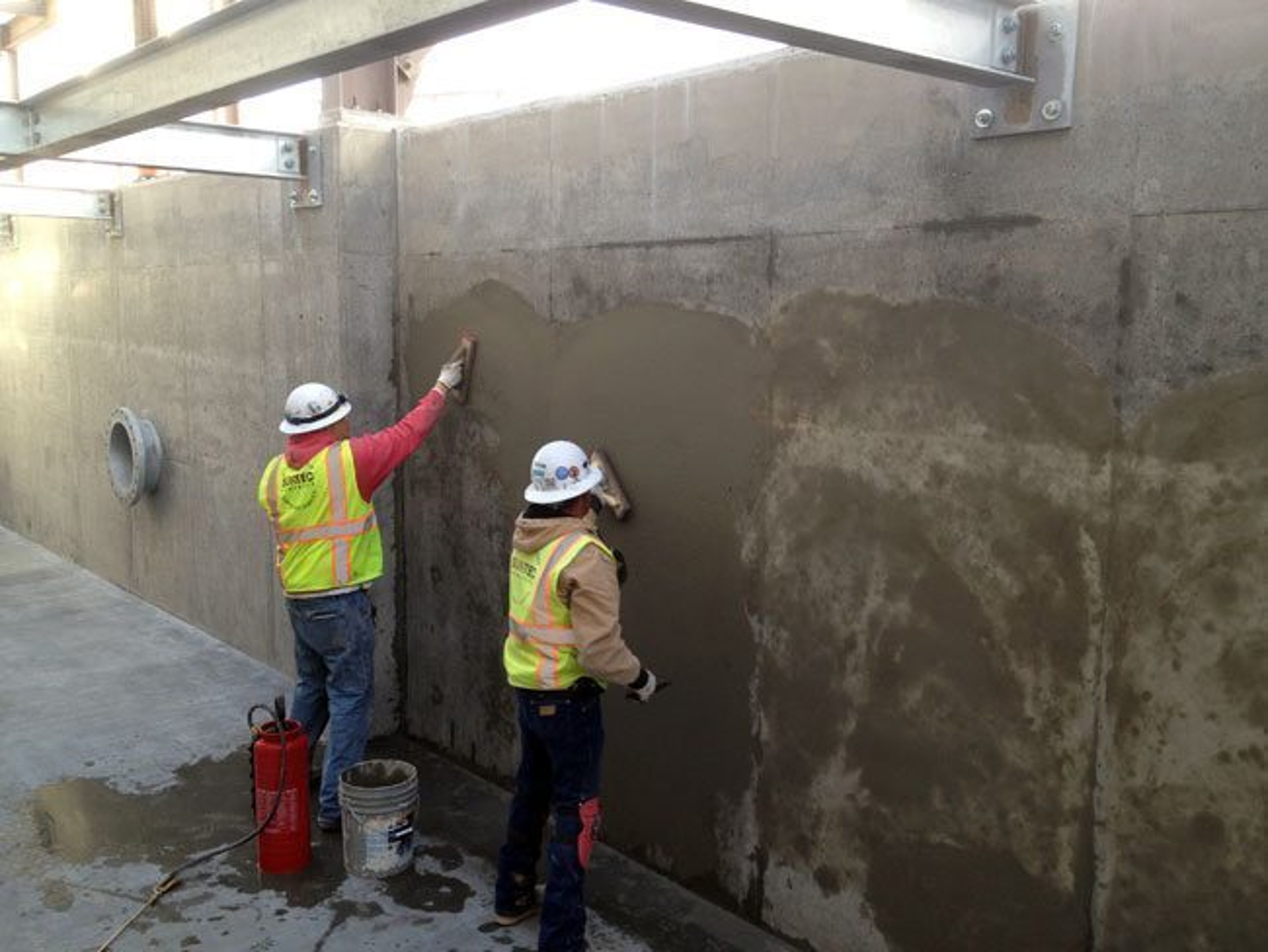 Two construction workers wearing safety vests and helmets are smoothing fresh concrete on a large wall with trowels. Construction equipment and water are on the ground nearby.