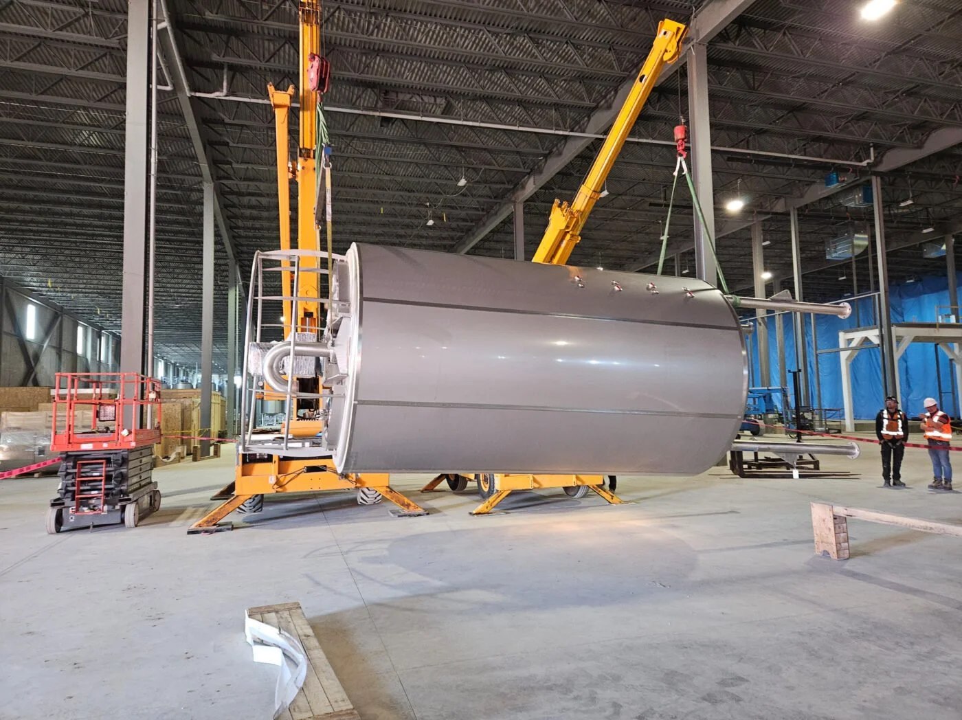 Large cylindrical metal component suspended by a crane inside a spacious industrial warehouse with workers observing.