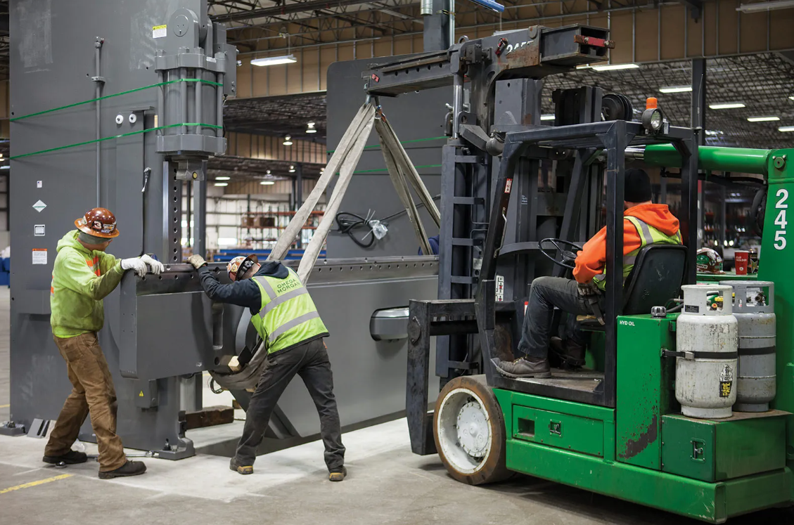 Workers assembling large industrial machinery inside a factory, with a forklift adjacent to the assembly.