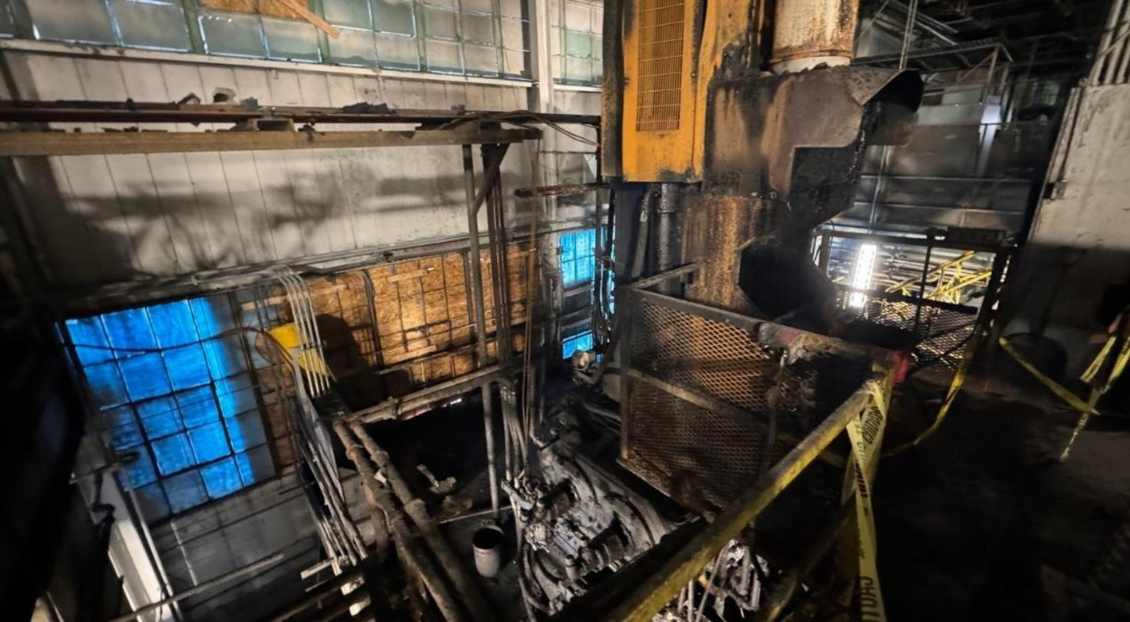 Industrial scene with machinery, pipes, and metal structures in a factory, showing signs of heat and soot. Yellow caution tape surrounds some equipment.
