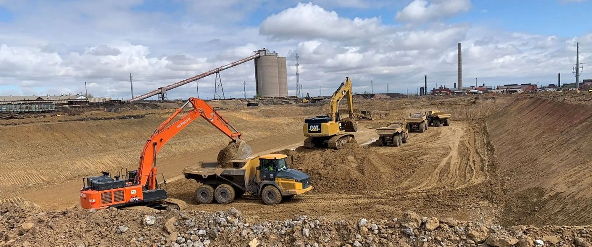 Construction site with excavators, dump trucks, and dirt mounds under a partly cloudy sky.