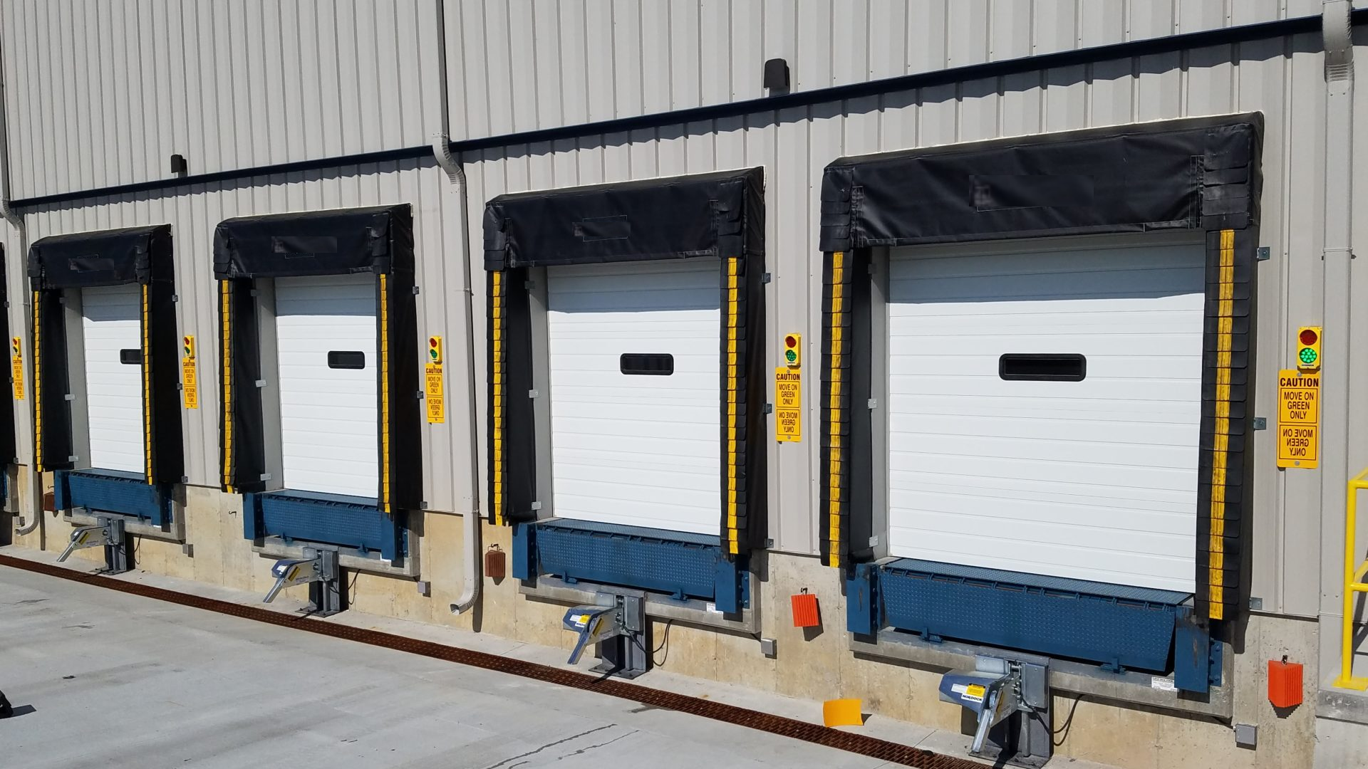 Four industrial loading dock doors with safety signs and markings on the exterior of a warehouse building.