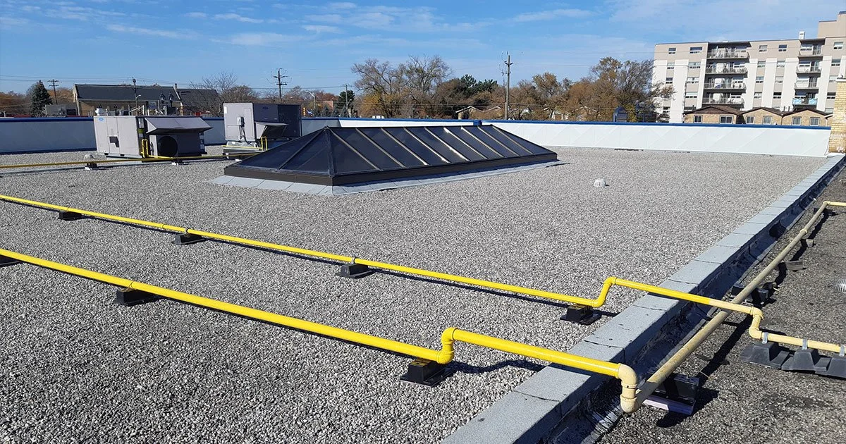 Flat rooftop with a vented skylight, yellow gas lines, and HVAC units in the background under a clear blue sky.