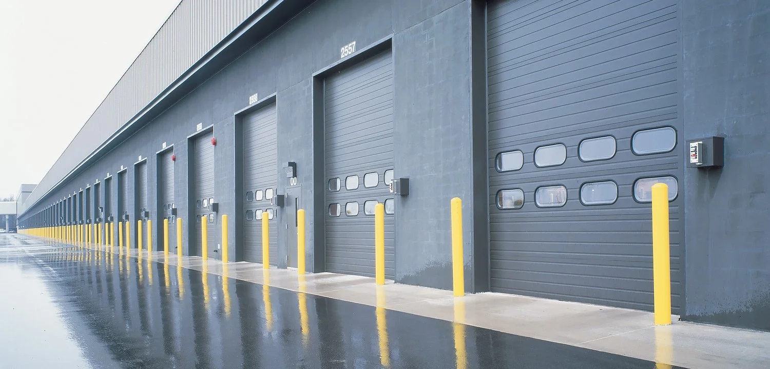 Exterior view of a row of warehouse loading docks with yellow safety bollards, gray rolling garage doors, and a wet reflective surface in front