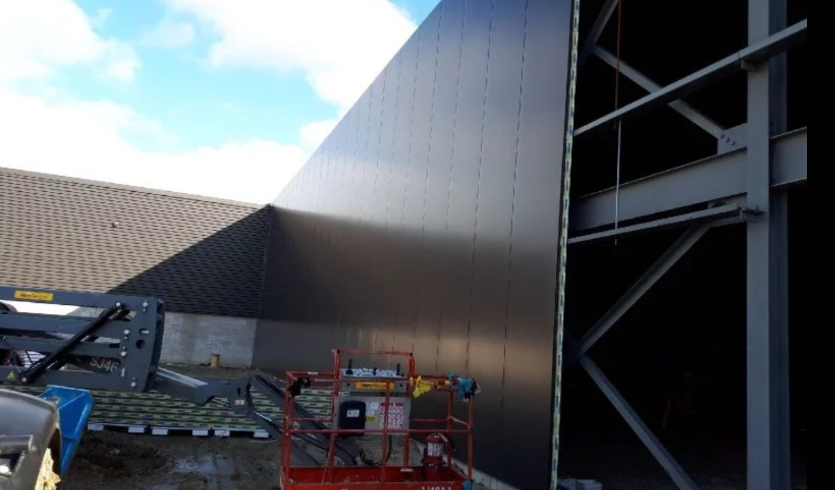Construction site with a large dark exterior wall and a partially built steel framework structure, equipment and a lift in the foreground.