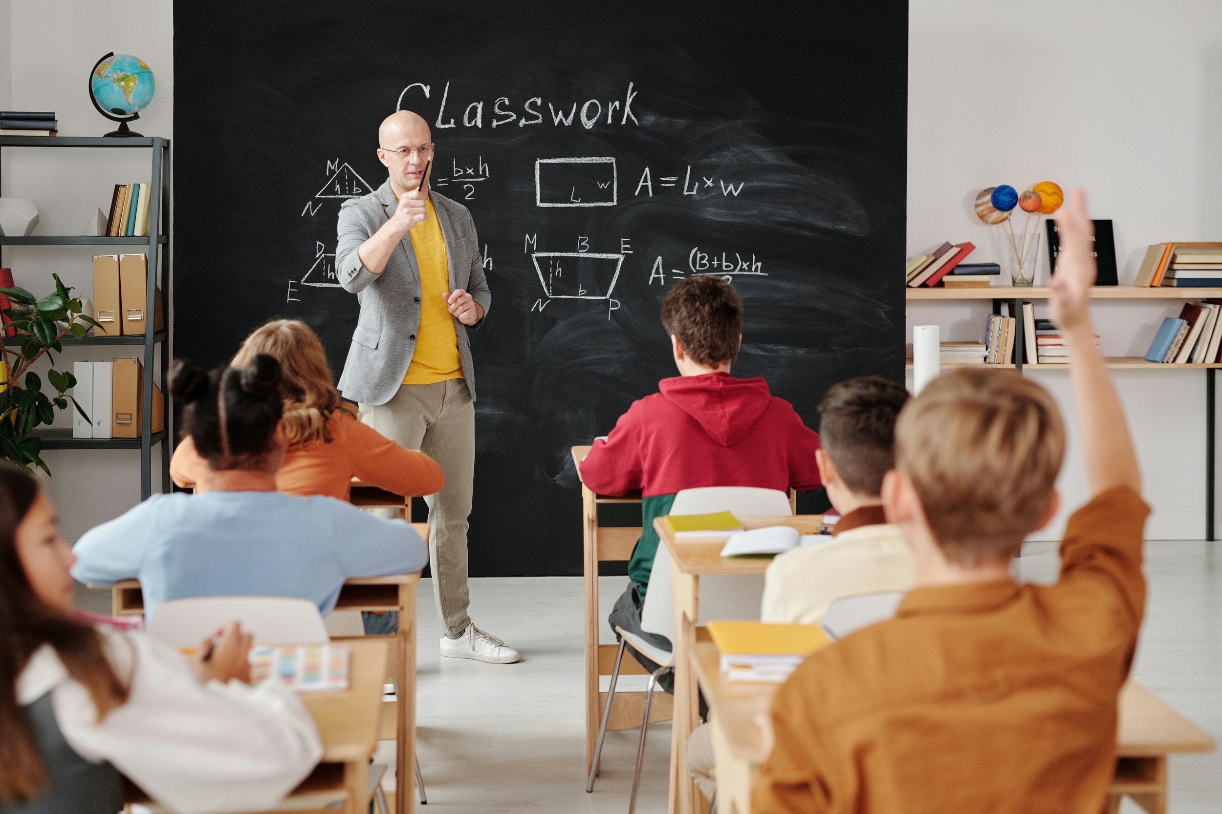 A classroom with a teacher at the blackboard and students raising their hands.