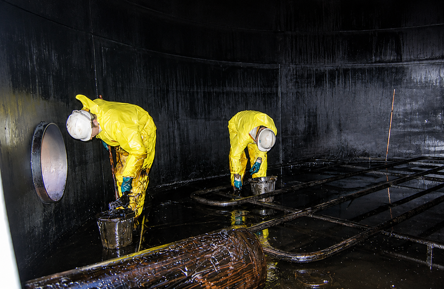 Two workers in yellow protective suits and helmets applying waterproofing or sealant to a black surface inside a large industrial tank or container.