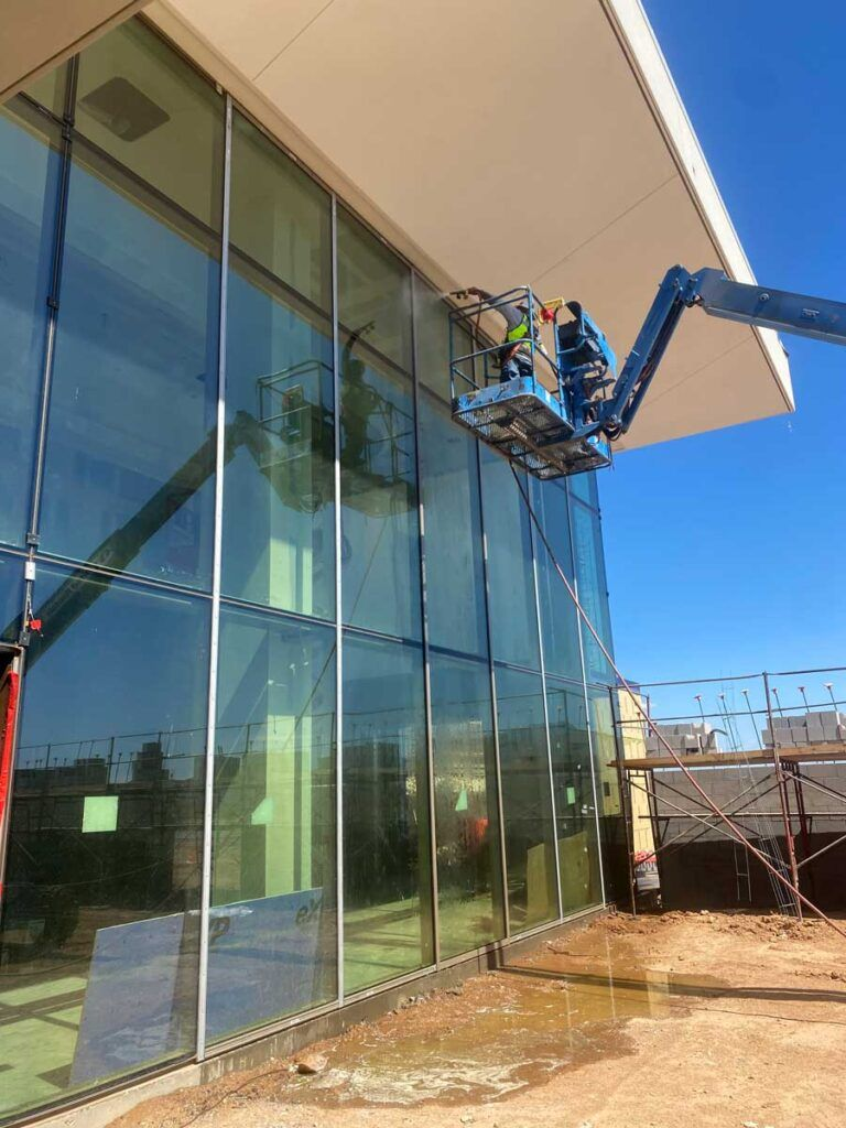 Construction workers on a lift cleaning or installing glass windows on a modern building with a white overhang and a clear blue sky.
