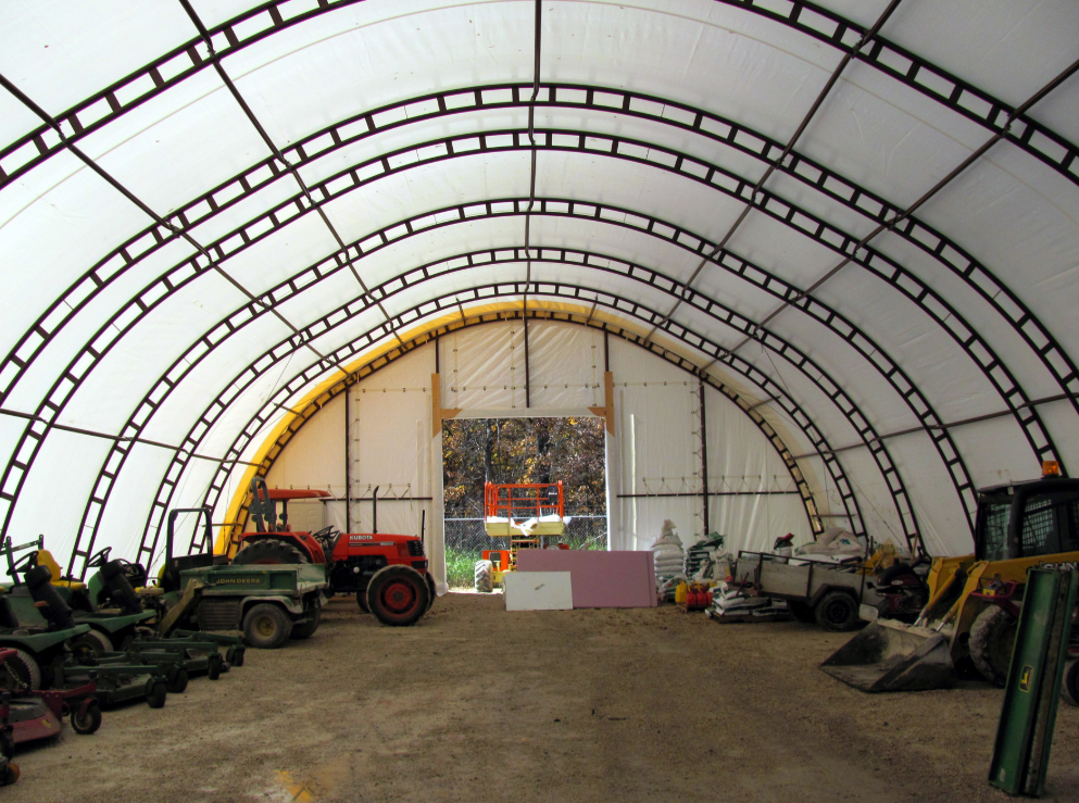 Inside a large, semi-circular greenhouse or storage tent with farming equipment, including tractors and lawnmowers, and various supplies and tools stored along the sides.
