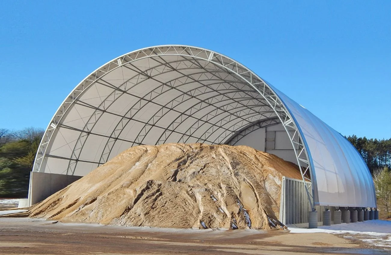 A large indoor structure with a curved white metal frame and translucent roof, housing a huge mountain of dirt or sand.