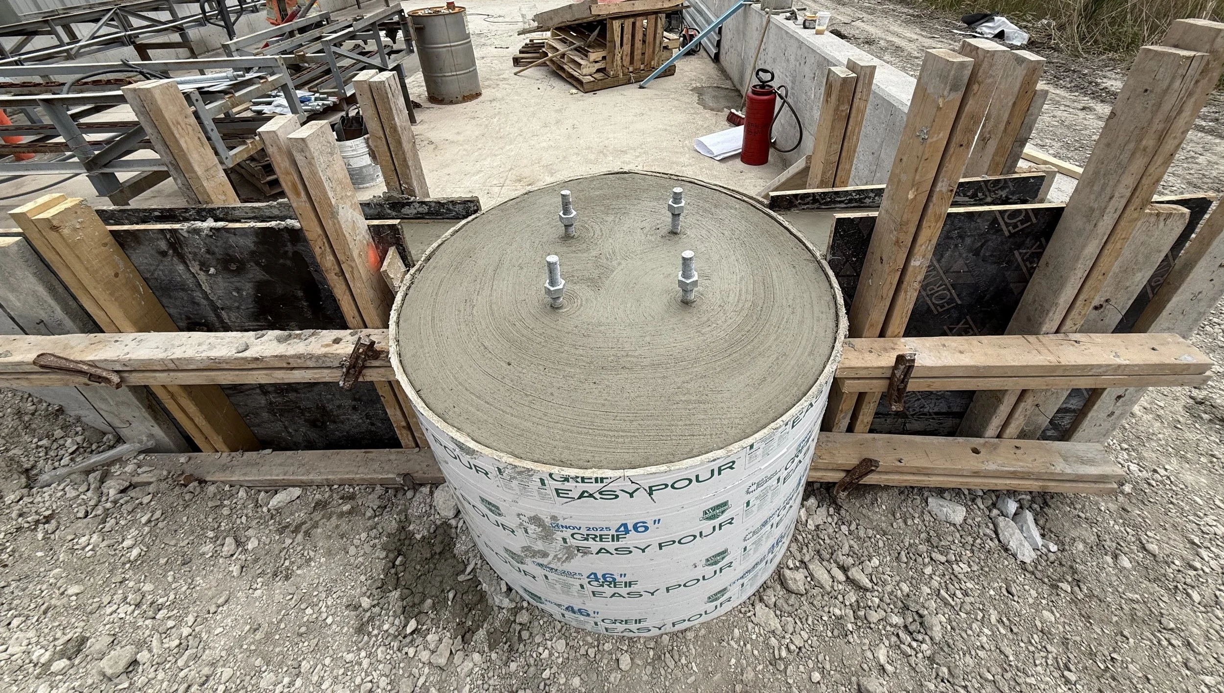 Construction site with a large concrete form tube and wooden framing for a poured concrete post or support.