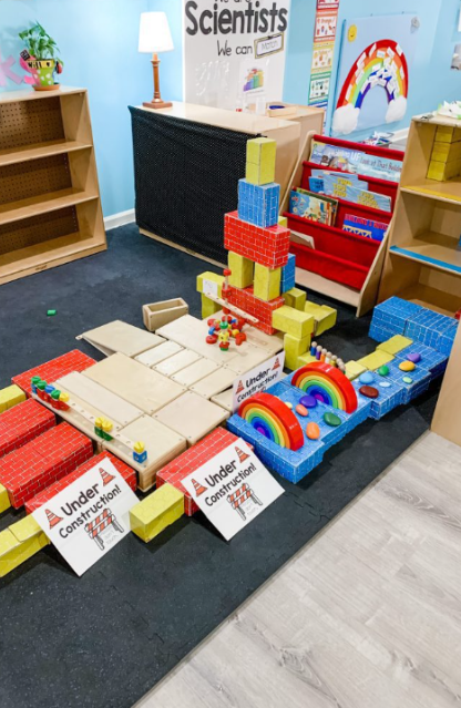 Large building block structure in a classroom under construction, with signs indicating under construction, surrounded by additional building blocks and toys.
