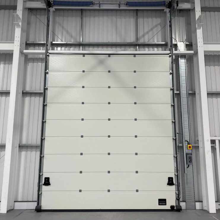 A large, white industrial garage door in a warehouse with metal framing and an emergency stop button on the right side.