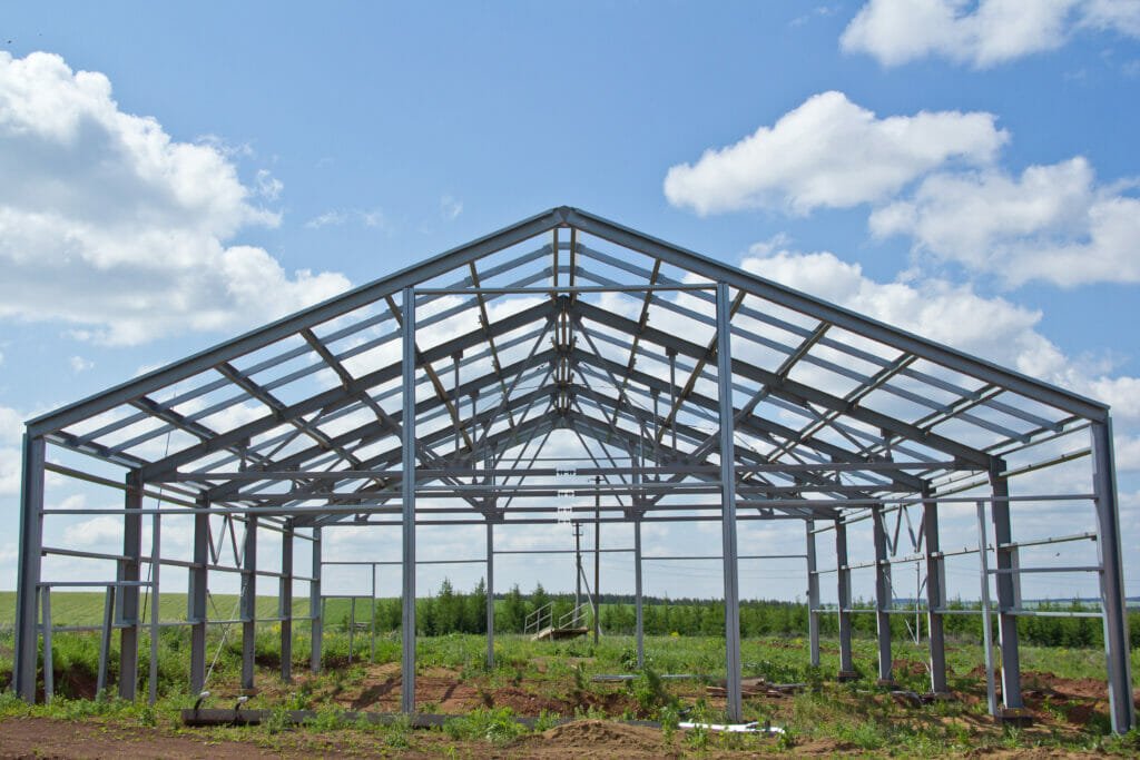 Steel frame structure of a greenhouse under construction, set on a grassy field with a blue sky and scattered clouds in the background.