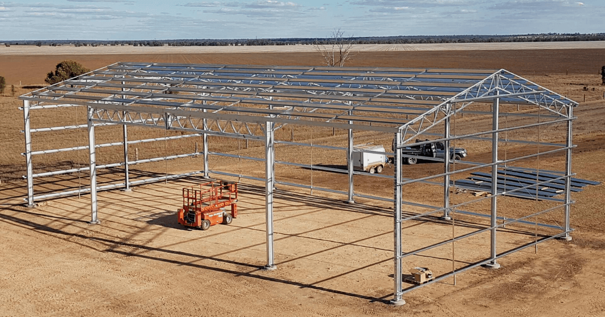 Steel frame structure being assembled for a building or greenhouse in a rural area, with construction equipment and vehicles around.