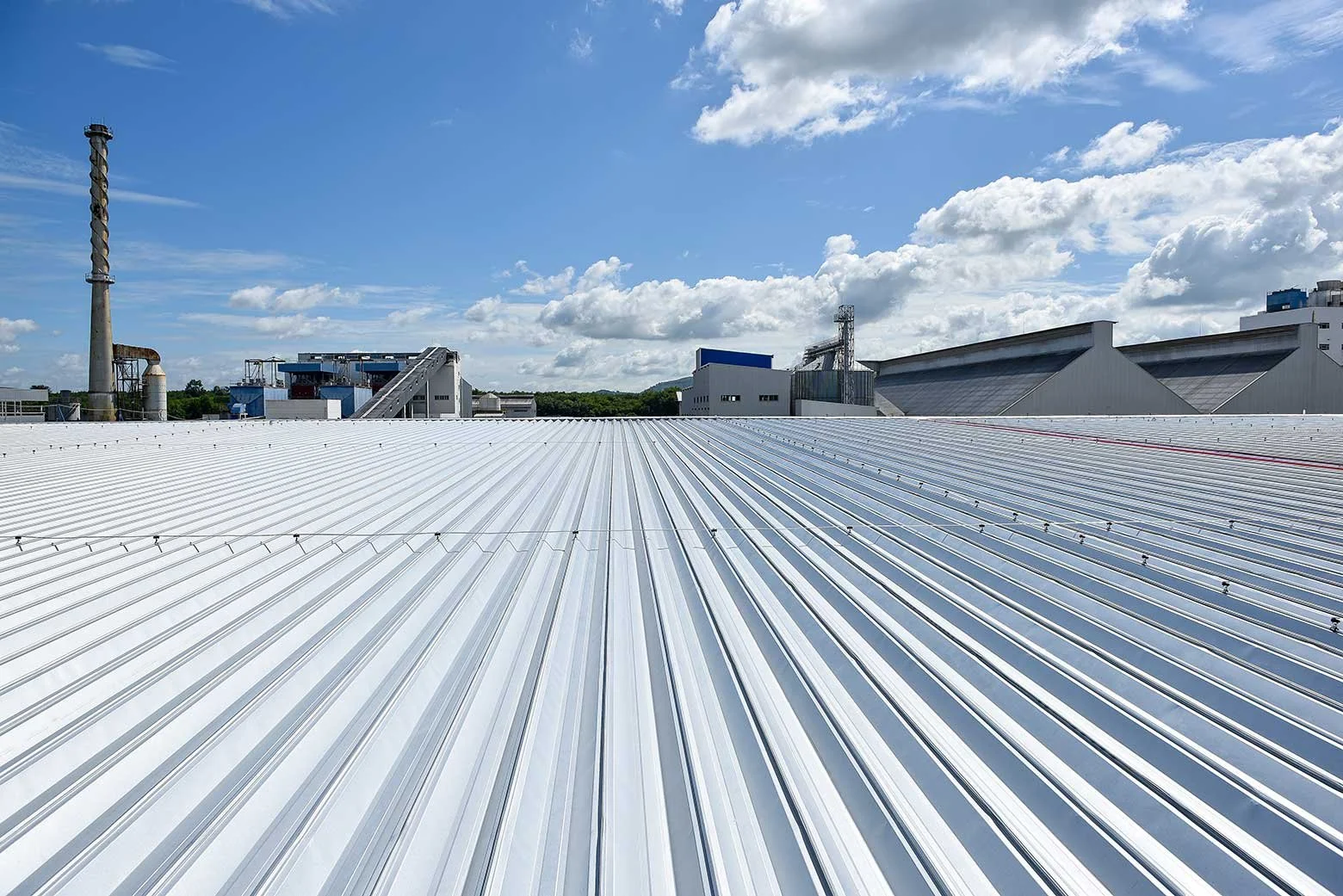 Large industrial building with a white metal roof, factory structures, and chimneys under a partly cloudy sky.