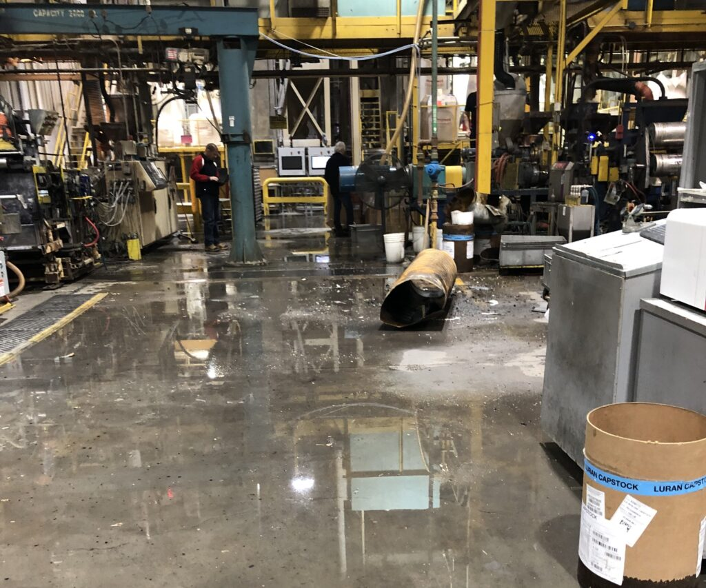 Industrial factory floor with machinery, workers, and scattered tools; flooded, wet floor with puddles and some spilled liquids.