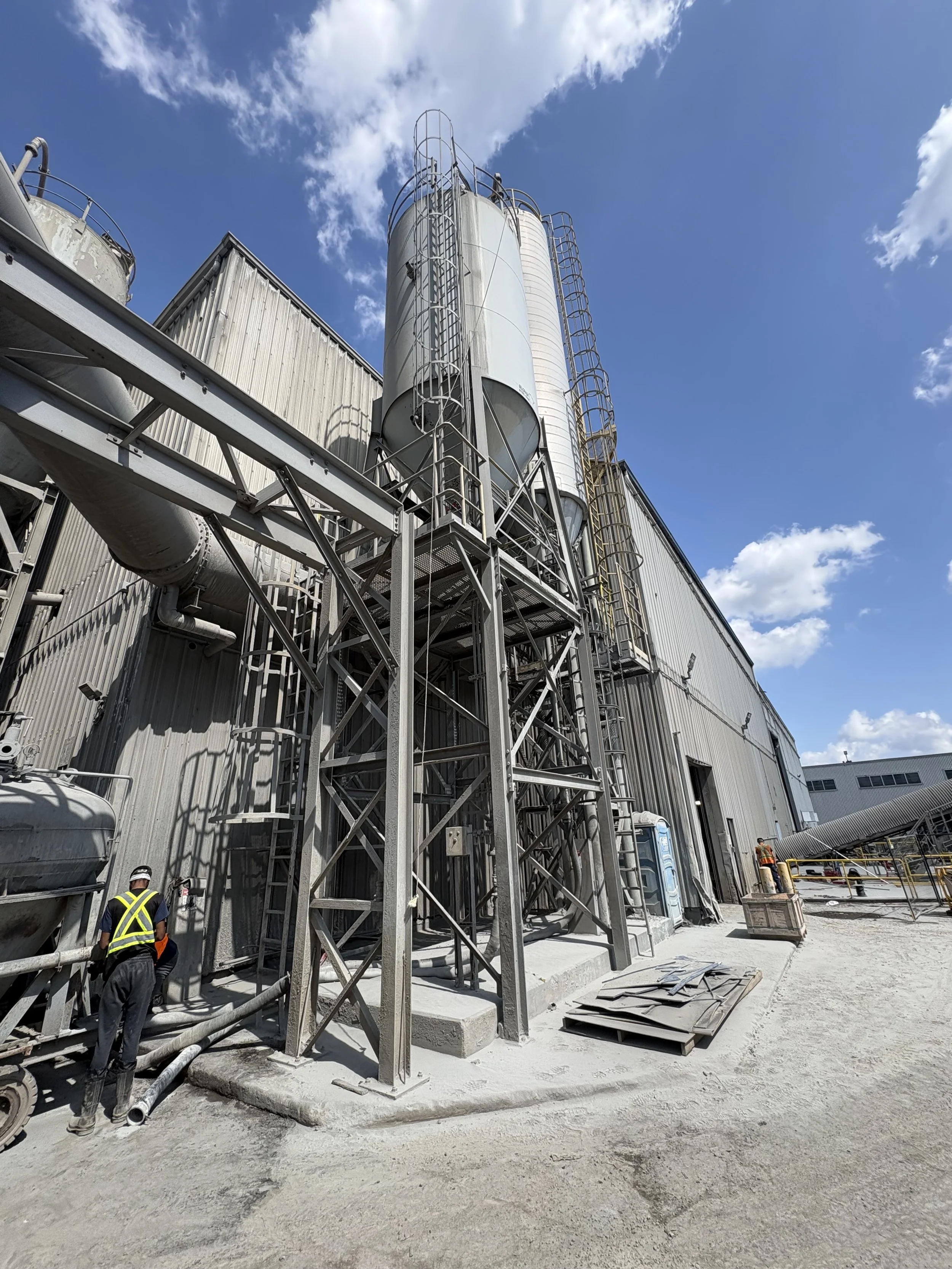 Construction workers at an industrial site, with large metal silos, pipes, and structural frameworks under a partly cloudy sky.