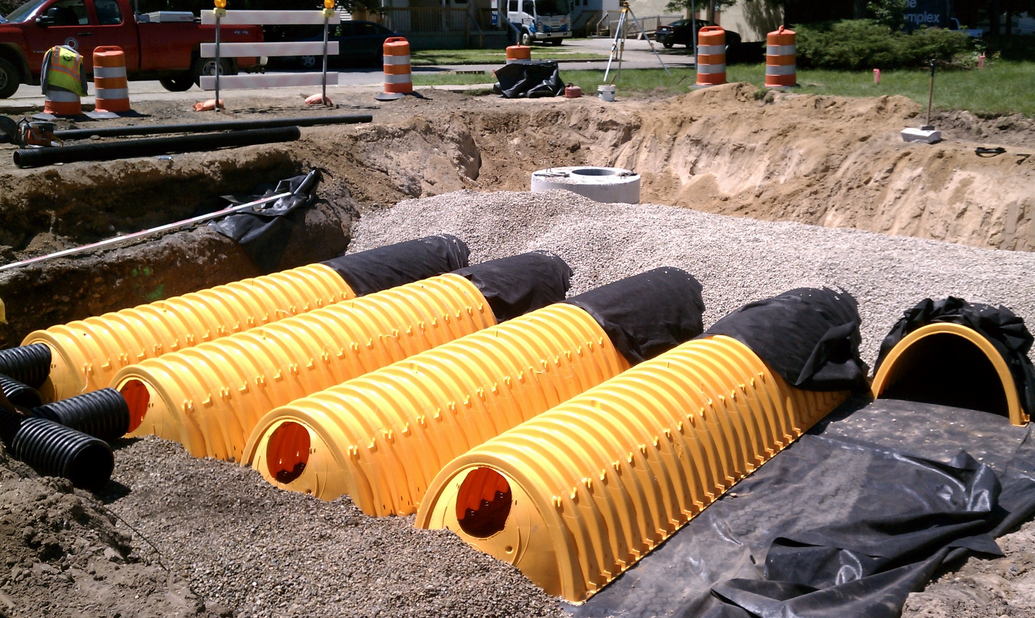 Construction site showing large yellow underground utility pipes being installed in a trench, with safety cones and equipment in the background.