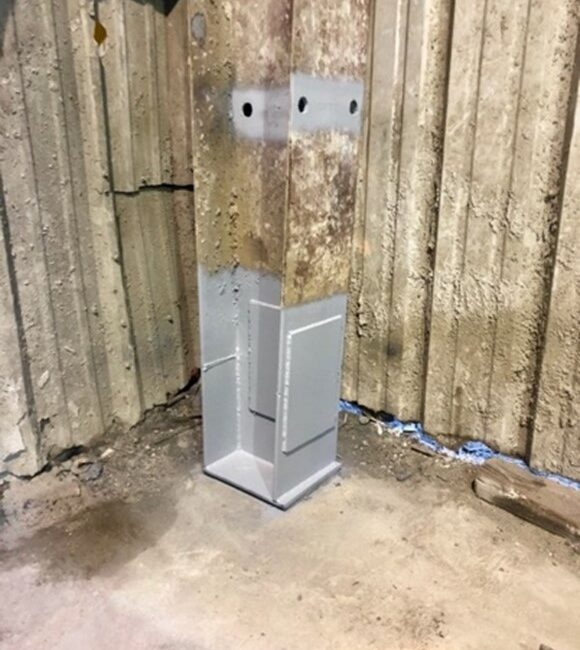 A metal electrical box installed in a wall with unfinished wood behind it, on a concrete floor.