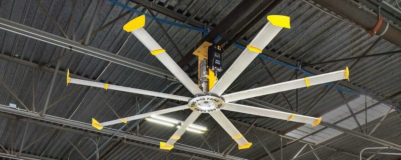 Large industrial ceiling fan with multiple white blades tipped with yellow, installed on a metal ceiling structure in what appears to be a warehouse or factory setting.