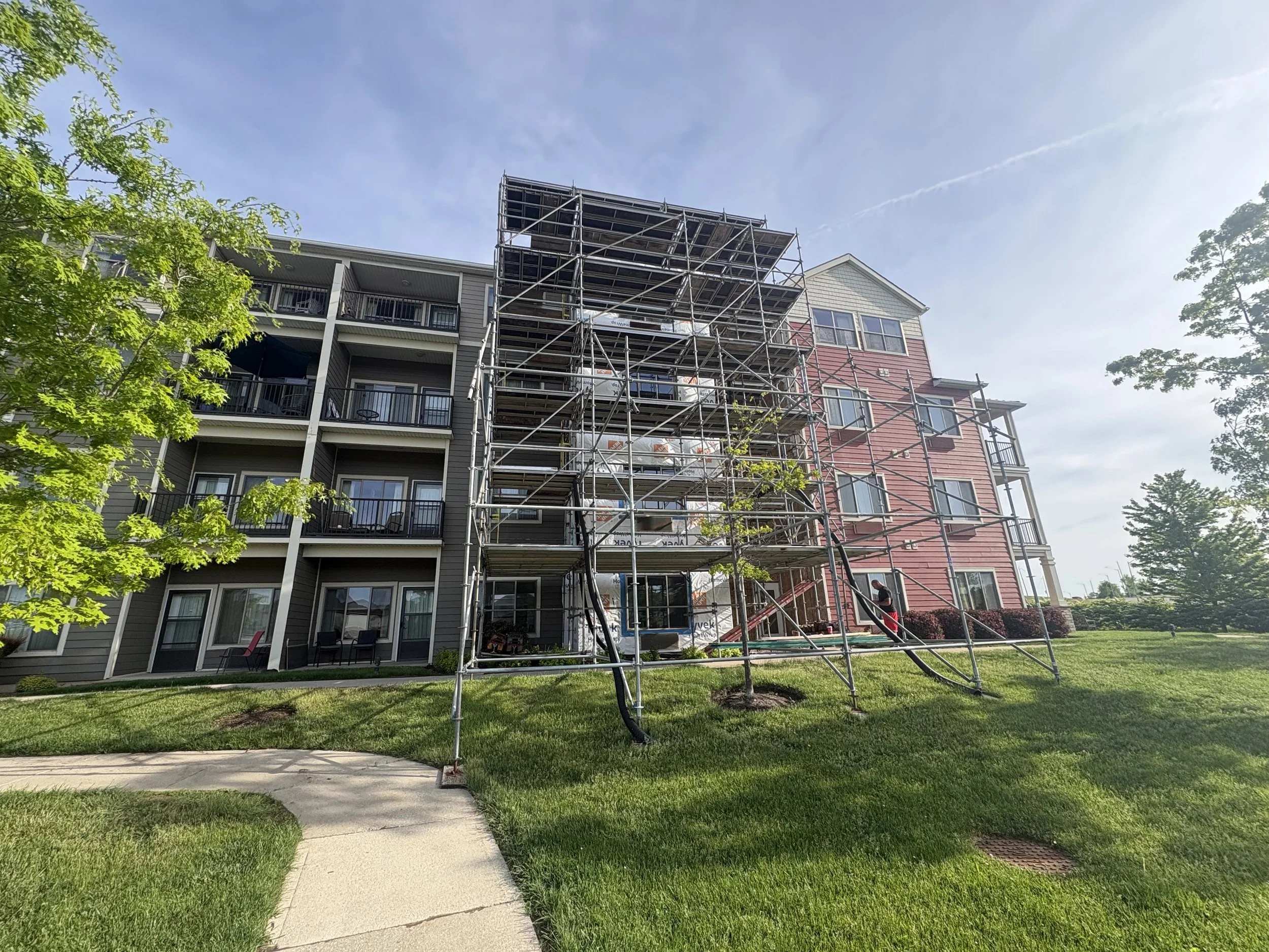 Multi-story residential building with scaffolding on the facade, surrounded by green trees and a lawn, under a blue sky with some clouds.
