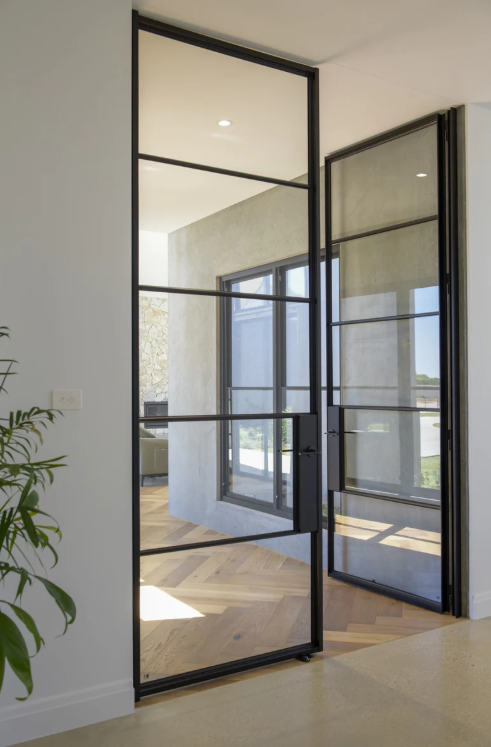 Interior view of a modern home with glass and metal framed doors leading to an outdoor space, natural light, light-colored walls, and wooden flooring.