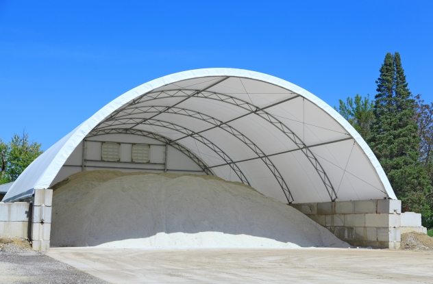A large outdoor storage tent filled with white granular material, with a blue sky and trees in the background.
