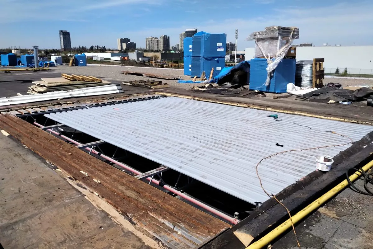 Construction site roof with a partially installed metal roof panel, construction materials, and blue equipment in the background with a city skyline.