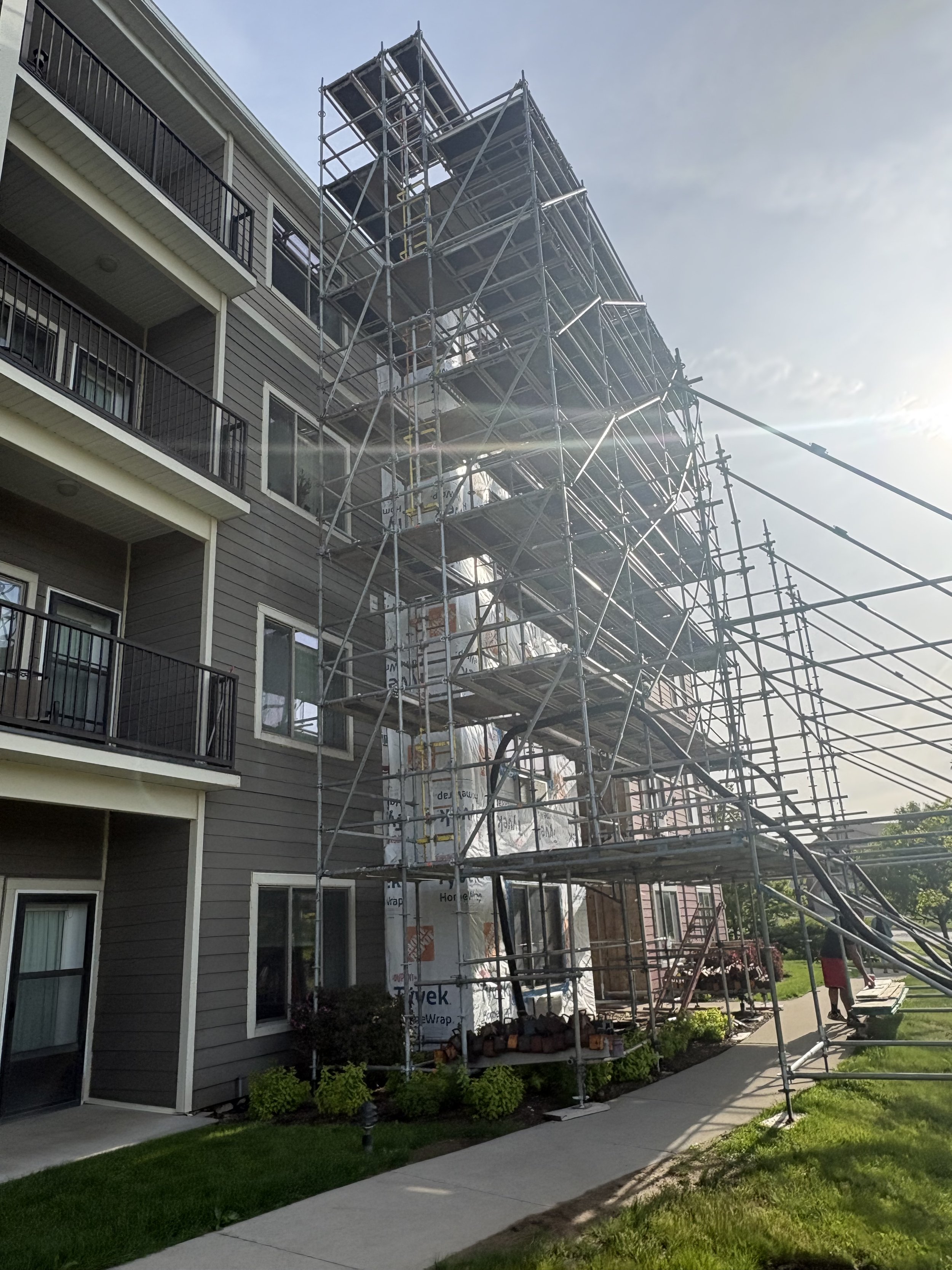 Apartment building with scaffolding set up on the right side, with construction materials stored underneath, and a manicured lawn and sidewalk in the front.