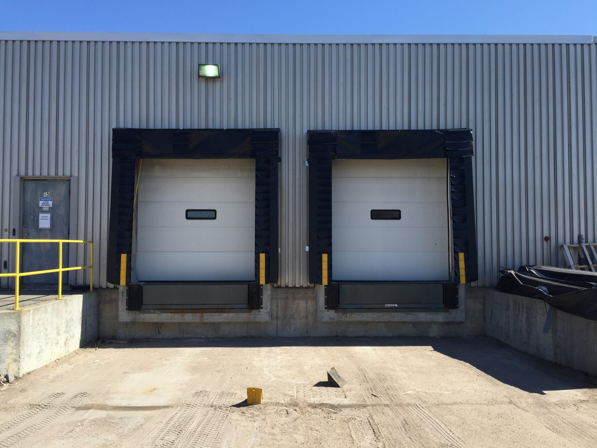 Two loading dock doors with black bumpers on a metal building exterior. A small door with a yellow handrail is on the left. The ground is dirt with tire tracks. There is a green light above the doors and a blue sky in the background.